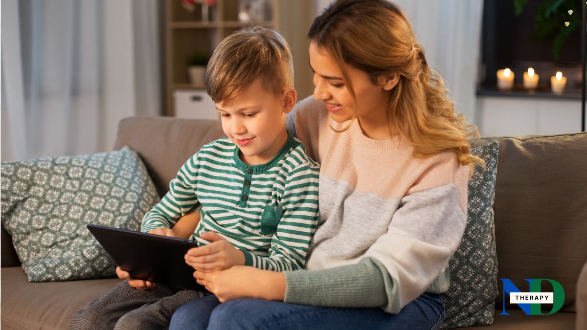 A mother and her young son sit together on a couch, calmly looking at a tablet, as she points to something on the screen.