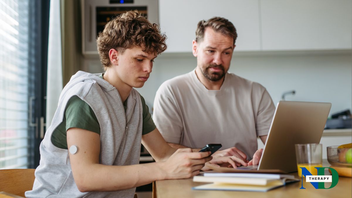 A parent and teenager sitting together at a kitchen table looking screens.